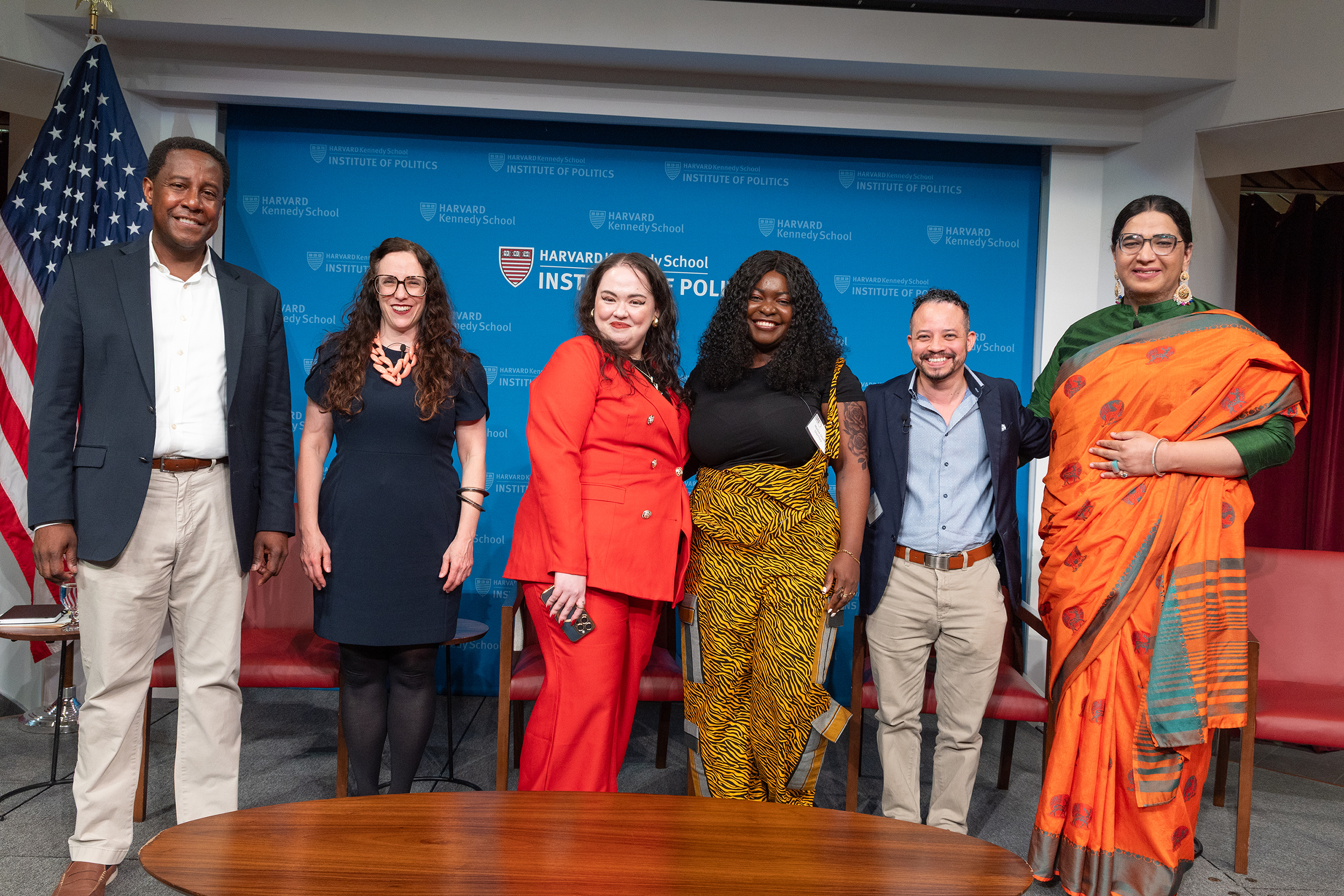 Six people stand side by side, smiling, in front of a Harvard Kennedy School Institute of Politics backdrop and an American flag.