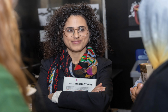 A woman with curly hair and glasses smiling, wearing a scarf and a badge, engaging in conversation with others at an event.