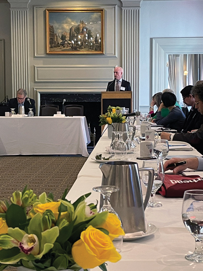 A speaker addresses an audience from a podium in a conference room with attendees sitting at tables adorned with flowers and water pitchers.