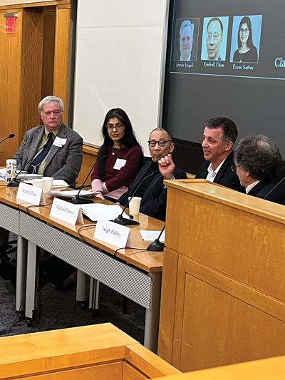 Five panelists at a conference table engaging in discussion, each with a nameplate, in a lecture hall setting.