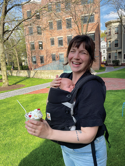 A woman smiling outdoors, with a baby in a front carrier, holding a cup of ice cream on a sunny day.