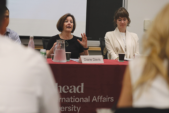 Two people sit at a panel table covered with a red cloth. A woman on the left speaks while gesturing, and a woman on the right listens with hands clasped. A name sign reads "Diane Davis.