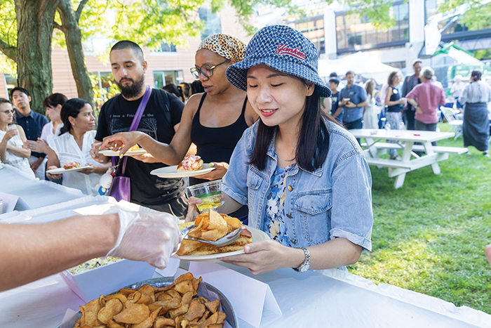 People serving themselves food outdoors at a buffet table with snacks and drinks, while others wait in line under the trees.