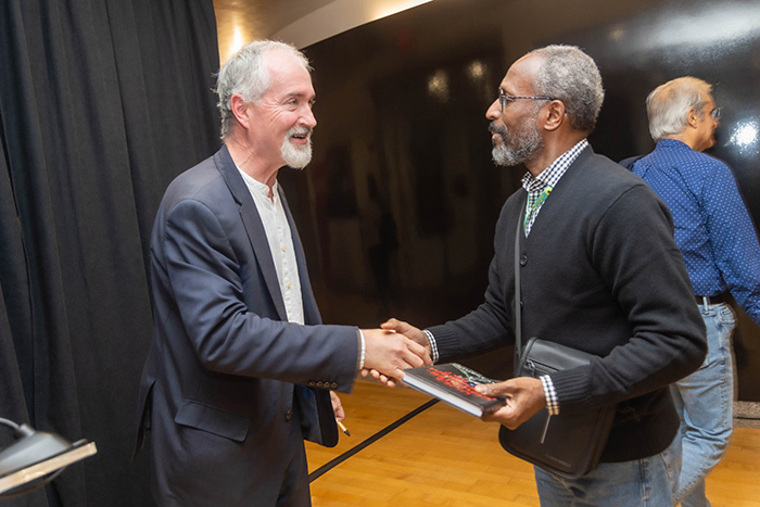 Two men shaking hands and smiling at each other in an indoor setting. One holds a book. Another person is in the background.