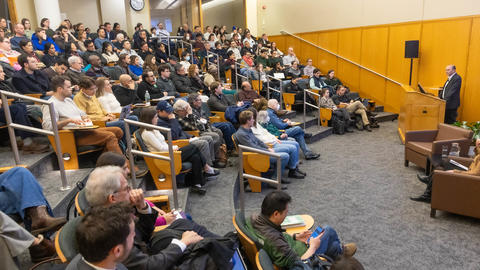 A large audience is listening to a man speak at a podium.