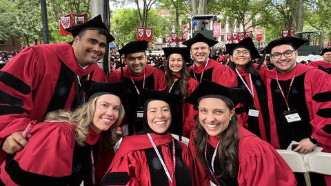 Nine graduate students in their red and black caps and gowns pose at graduation ceremony.