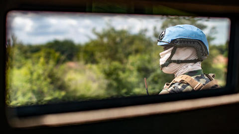 Profile of man with army fatigues and face covered wearing a blue helmet is seen through a car window.