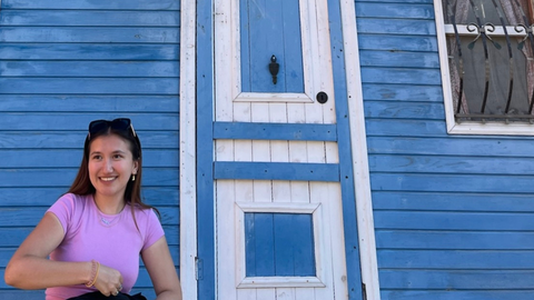 A smiling woman stands in front of a blue wooden building with white trim.
