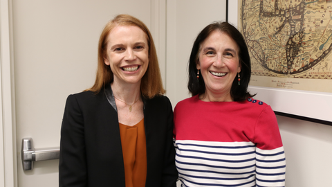 Two women standing indoors, smiling at the camera; one wears a black blazer and orange top, the other wears a red and white striped shirt, with a framed map on the wall behind them.