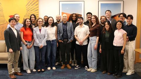 A group of 19 people pose and smile for a photo in an office setting, standing in front of sofas and framed artwork on the wall.