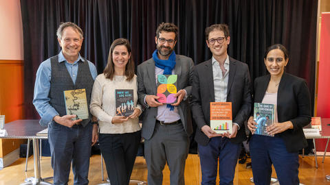 Five people standing indoors, each holding a book, with a black curtain and tables in the background. 