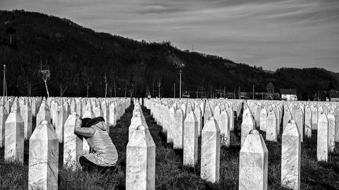 Woman kneels in front of a white gravestone in a large cemetery filled with identical gravestones. 
