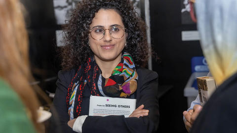 A woman with curly hair and glasses smiling, wearing a scarf and a badge, engaging in conversation with others at an event.