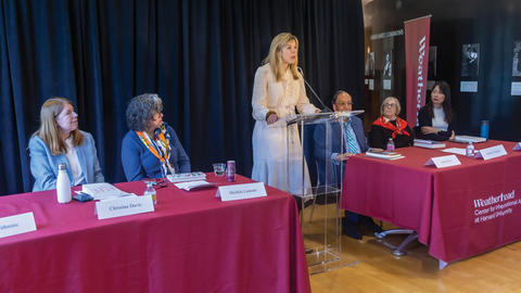 A woman speaks at a podium during a panel discussion with four seated panelists in a conference room at a university.