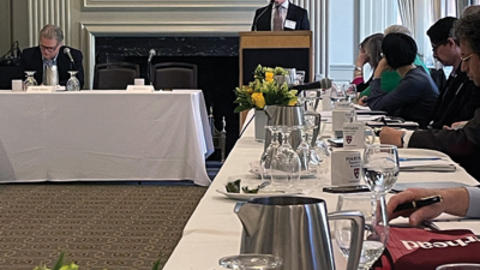 A speaker addresses an audience from a podium in a conference room with attendees sitting at tables adorned with flowers and water pitchers.