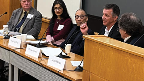 Five panelists at a conference table engaging in discussion, each with a nameplate, in a lecture hall setting.