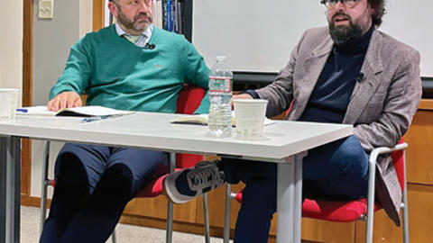Two men sitting at a table in a library, engaged in a discussion, with bookshelves in the background.