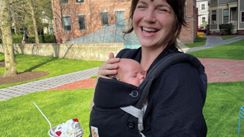 A woman smiling outdoors, with a baby in a front carrier, holding a cup of ice cream on a sunny day.