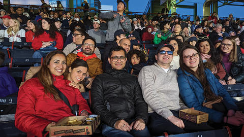 A group of affiliates enjoying a baseball game from stadium seats, smiling at the camera, with other spectators and the field in the background.