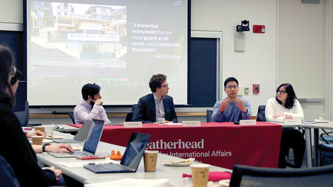 Four people sitting at a conference table engaging in a discussion, with an audience and a large projector screen in the background displaying a presentation.