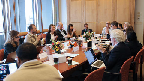 A group of professionals clapping while sitting around a conference table, engaged in a meeting, with papers and laptops visible.