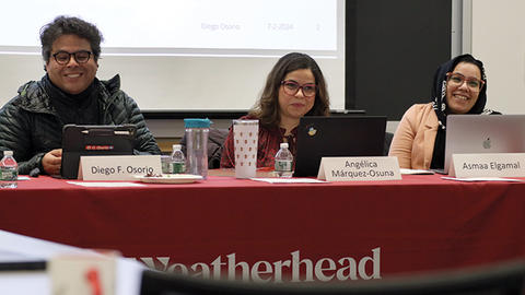 Three smiling academics sit at a table with laptops during a conference, with name tags visible and a screen in the background.