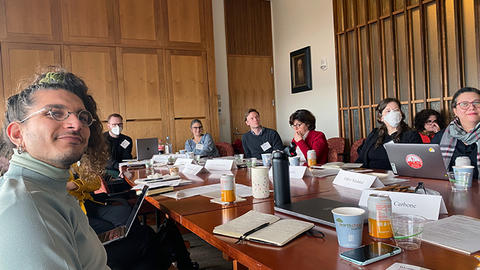 A group of diverse professionals sitting around a conference table, engaged in a meeting, with papers and laptops visible.