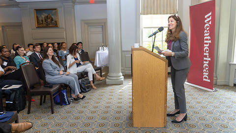 A woman speaks at a podium to an audience in a formal, carpeted room. A banner with "Weatherhead" is visible in the background.