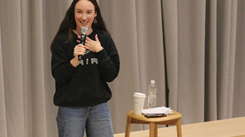 A woman speaking into a microphone on stage, standing next to a stool with a water bottle and cup.
