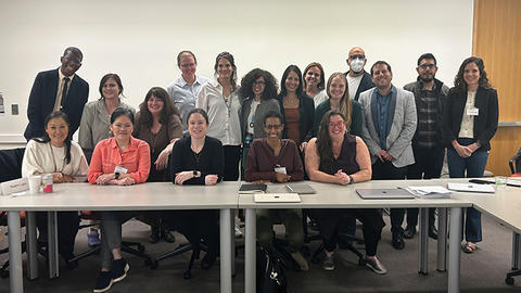 A group of people smiling, standing and sitting behind a conference table in a classroom setting.