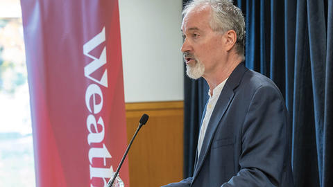A man in a suit speaks at a podium with a microphone. A red banner with partial text "Weath" is visible in the background.