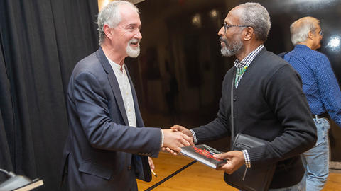 Two men shaking hands and smiling at each other in an indoor setting. One holds a book. Another person is in the background.
