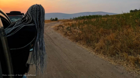 A woman with long dyed braids is leaning out of a car and looking down the road toward a sunset.