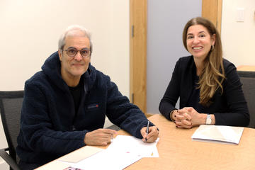 An older man sits at a table signing papers with a pen, while a woman sits next to him smiling. Both are indoors at a desk.