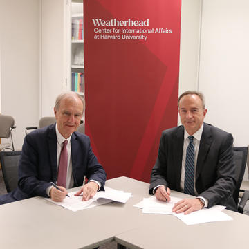 Two men in suits sit at a table, signing documents, with a Weatherhead Center for International Affairs at Harvard University banner in the background.
