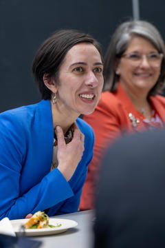 Two women sit at a table, engaged in conversation; the woman in the foreground wears a blue blazer and is smiling, while the woman in the background wears an orange blazer.