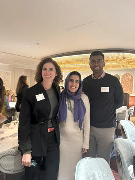 Three people stand together and smile for a photo at an indoor event. Two women and one man wear name tags and business casual attire. Dining tables and other attendees are visible in the background.