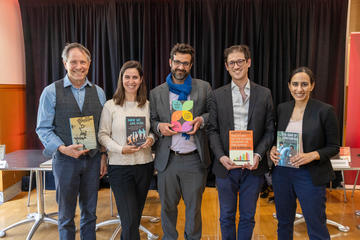 Five people standing indoors, each holding a book, with a black curtain and tables in the background. 