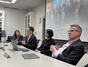 Four panelists sit at a conference table during a discussion on the Dayton Peace Agreement, with a presentation slide visible on the screen behind them. 