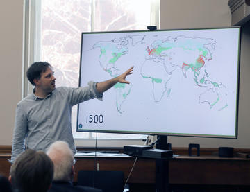 A man gestures toward a world map displayed on a screen, highlighting regions in color with the year 1500 shown, while an audience watches in a classroom setting. 