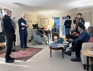 A group of people are gathered in a living room, some seated and others standing, engaged in conversation and holding papers. 