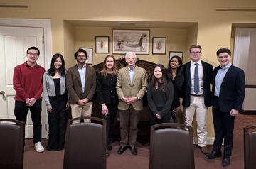 Eight people stand together, posing for a group photo in a dining room with beige walls and framed art behind them. Some are dressed in business attire. 
