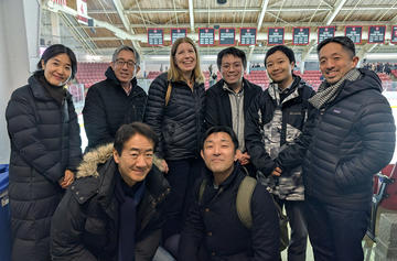A group of eight adults wearing winter jackets pose together inside an indoor ice rink with bleachers and banners in the background. 