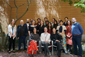 A group of 20 people pose for a photo outdoors in front of a terracotta wall, with some seated and others standing. 