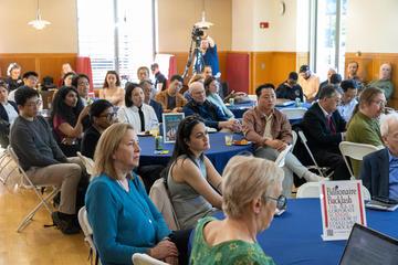 A diverse group of people sit at round tables, listening attentively at an indoor event; one table displays books and signs, and a cameraman films in the background.