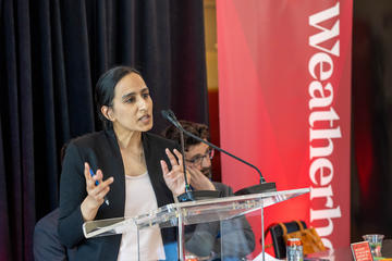 A woman speaks at a clear podium with a microphone, gesturing with her hands. A crimson Weatherhead banner is visible in the background.
