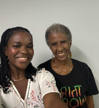 Two women smiling indoors, one wearing a floral blouse and the other a black t-shirt with colorful text.