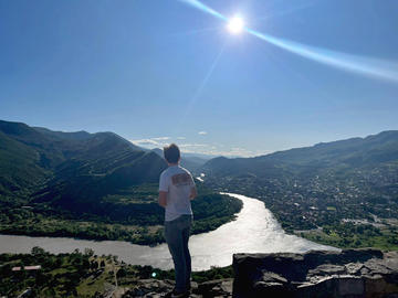 A person stands on a rocky ledge, facing a winding river that cuts through a valley under a clear blue sky with the sun shining brightly.