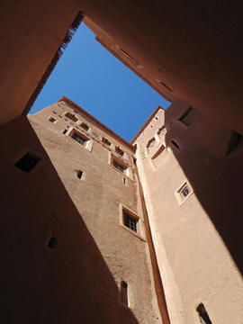 View looking up from a narrow alley between tall sandstone buildings with small windows, showcasing a slice of blue sky.