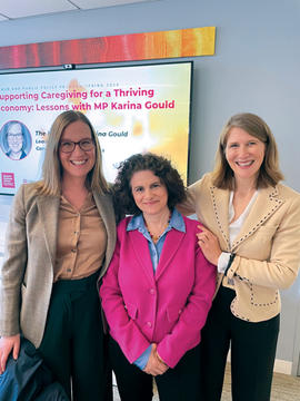 Three women smiling and standing together in front of a presentation screen with text about caregiving and economy discussions.